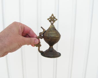 May include: An antique brass incense burner with intricate floral patterns and a decorative handle. The burner features a cross-shaped finial on top and a pedestal base. The overall design is ornate and detailed.