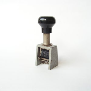 Könnte beinhalten: Ein vintage Zahlen-Stempel aus silberfarbenem Metall mit einem schwarzen Kunststoffgriff. Der Stempel hat eine Reihe von drehbaren Rädern mit Zahlen darauf.