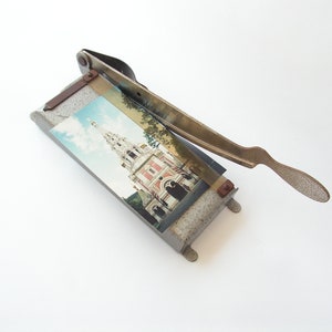 May include: A vintage metal photo cutter with a gray base and a metal handle. The cutter has a photo of a building with a spire on it.