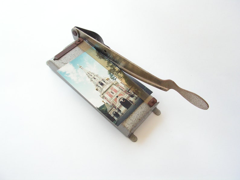 May include: A vintage metal photo cutter with a gray base and a metal handle. The cutter has a photo of a building with a spire on it.