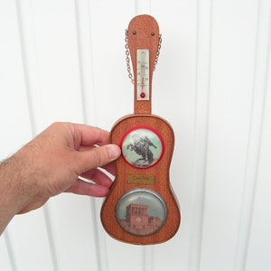 May include: A wooden guitar-shaped thermometer with a red and white thermometer scale, a red knob, and a chain. The guitar has two round images, one of a man on horseback and the other of a building. The text "Espeluz" is on the guitar.
