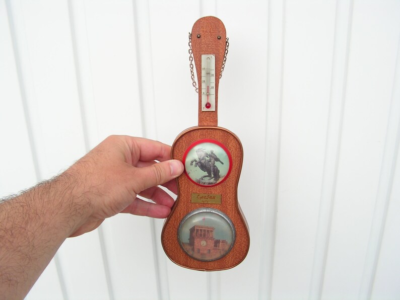 May include: A wooden guitar-shaped thermometer with a red and white thermometer scale, a red knob, and a chain. The guitar has two round images, one of a man on horseback and the other of a building. The text "Espeluz" is on the guitar.
