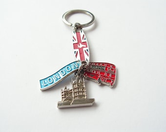 May include: A silver keychain with a blue and white ribbon that says "LONDON" and features a red double-decker bus, a silver replica of the Tower Bridge, and a red, white, and blue enamel charm with the Union Jack flag.