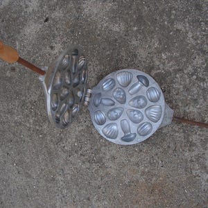 May include: Open, vintage metal cookie mold with a variety of shell and mushroom shapes. The mold is silver with a wooden handle and is open, revealing the detailed impressions for baking. The handle is brown.