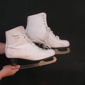 May include: A pair of white figure skates with black laces and a black inscription that reads "Dagmar by sien." The skates have a gold blade.