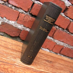 May include: A vintage, dark brown book titled "Dictionnaire Quillet de la Langue Fran&ccedil;aise" is propped against a red brick wall. The book's spine displays gold lettering and the phrase "L'art d'&eacute;crire et de bien r&eacute;diger". The book is resting on a wooden surface.