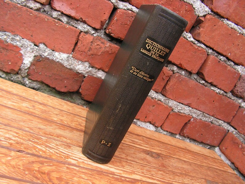 May include: A vintage, dark brown book titled "Dictionnaire Quillet de la Langue Fran&ccedil;aise" is propped against a red brick wall. The book's spine displays gold lettering and the phrase "L'art d'&eacute;crire et de bien r&eacute;diger". The book is resting on a wooden surface.