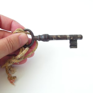 May include: An antique metal key with a circular handle and a rectangular bit. The key is attached to a beige and tan rope. The key shows signs of age and wear, with a rusty appearance.