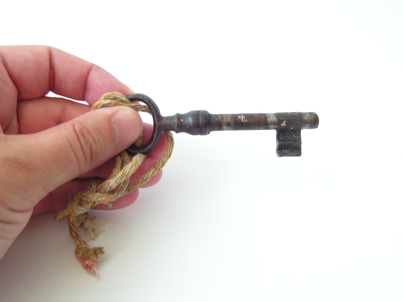 May include: An antique metal key with a circular handle and a rectangular bit. The key is attached to a beige and tan rope. The key shows signs of age and wear, with a rusty appearance.