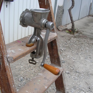 Può includere: Un macinaca carne manuale in metallo argentato con una manovella in legno, montato su una scala di legno.