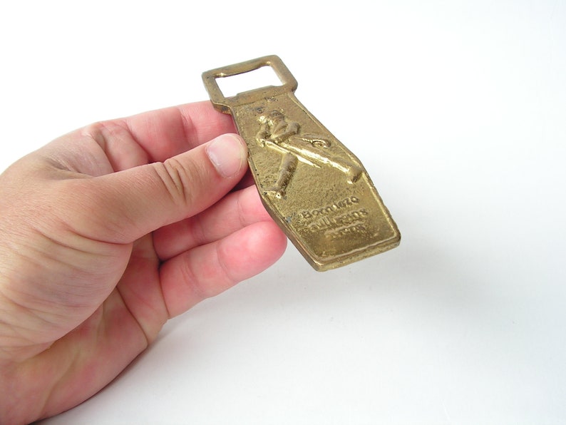 May include: A gold-coloured bottle opener with a raised image of a baseball player and the text "Boston Red Sox" on the front.