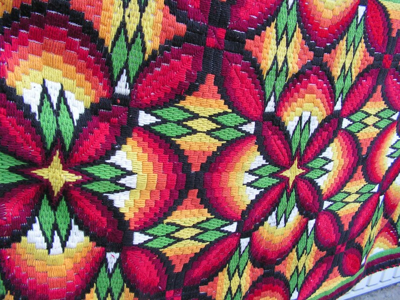 May include: Close-up of a colorful, embroidered textile with a repeating floral pattern. The design features red, orange, yellow, green, and black threads, creating a vibrant and textured surface. The textile appears to be a decorative piece.