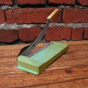 May include: A green paper cutter with a silver blade and a yellow handle. The cutter is sitting on a wooden surface.