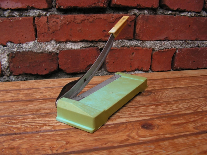 May include: A green paper cutter with a silver blade and a yellow handle. The cutter is sitting on a wooden surface.