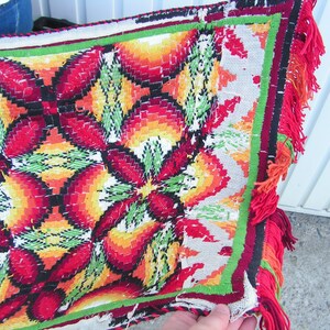 May include: A colorful, hand-stitched textile with a repeating floral pattern in red, orange, yellow, and green. The edges are trimmed with a red fringe. The textile appears to be a decorative piece, possibly a throw or wall hanging.