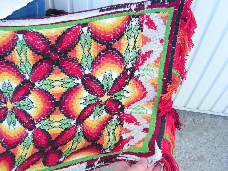 May include: A colorful, hand-stitched textile with a repeating floral pattern in red, orange, yellow, and green. The edges are trimmed with a red fringe. The textile appears to be a decorative piece, possibly a throw or wall hanging.