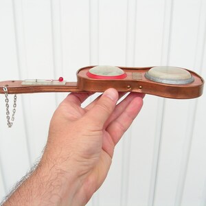 May include: A small, copper-colored, guitar-shaped object with two round, white, and silver-colored dials. The object has a small, red button on the top and a silver chain attached to the bottom.
