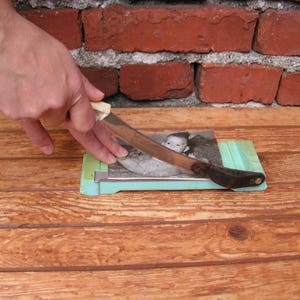 May include: A person is using a paper cutter to cut a black and white photograph of a baby. The photograph is on a light blue cutting mat. The person's hand is holding a silver knife with a white handle.