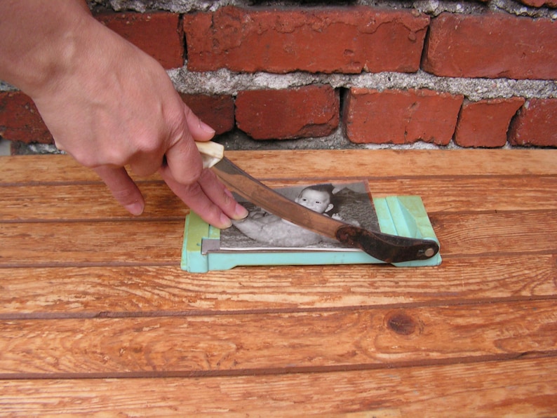 May include: A person is using a paper cutter to cut a black and white photograph of a baby. The photograph is on a light blue cutting mat. The person's hand is holding a silver knife with a white handle.