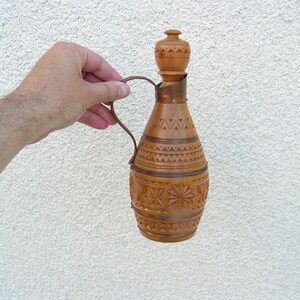 K&ouml;nnte beinhalten: Eine Holzflasche mit einem geschnitzten Design und einem kupfernen Griff. Die Flasche ist braun und hat einen hellbraunen Holzkopf.