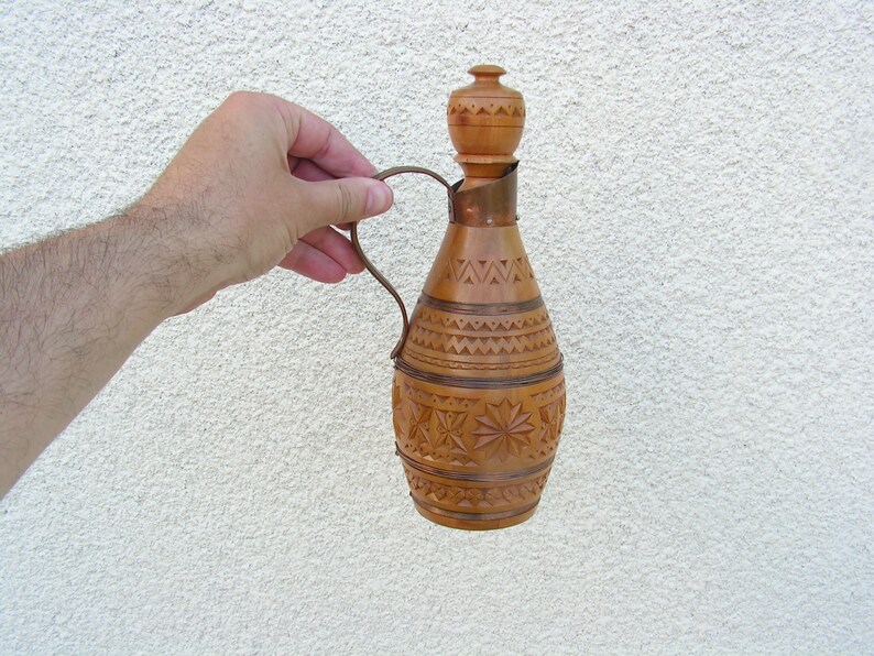 K&ouml;nnte beinhalten: Eine Holzflasche mit einem geschnitzten Design und einem kupfernen Griff. Die Flasche ist braun und hat einen hellbraunen Holzkopf.