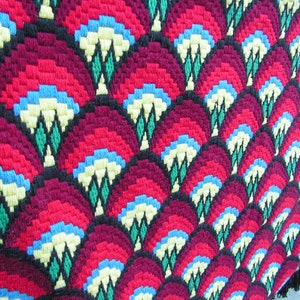 May include: Close-up of a colorful, patterned textile. The design features repeating fan-like shapes in shades of red and burgundy, with accents of blue, yellow, green, and black. The textile has a textured appearance, suggesting a woven or embroidered construction.