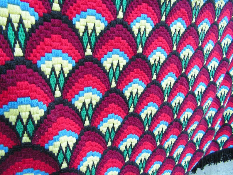 May include: Close-up of a colorful, patterned textile. The design features repeating fan-like shapes in shades of red and burgundy, with accents of blue, yellow, green, and black. The textile has a textured appearance, suggesting a woven or embroidered construction.
