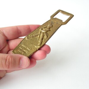 May include: A gold-coloured bottle opener with a raised design of a person holding a bottle. The opener has a simple, classic design.