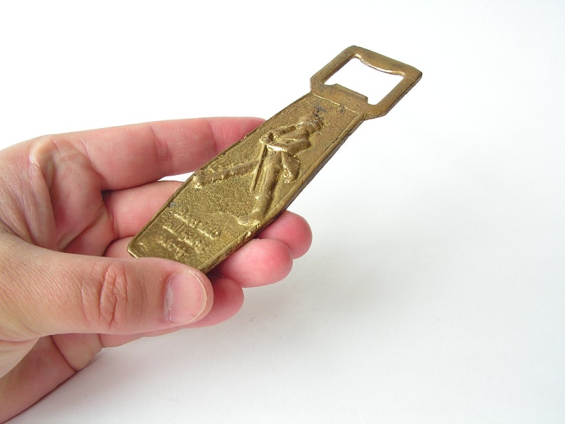 May include: A gold-coloured bottle opener with a raised design of a person holding a bottle. The opener has a simple, classic design.