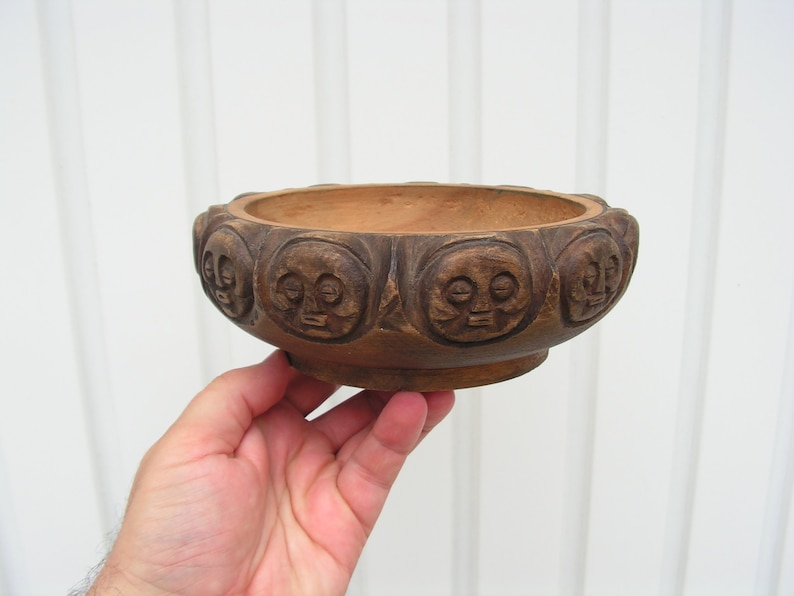 May include: A wooden bowl with a carved design of faces around the rim. The bowl is brown and has a smooth, polished finish.