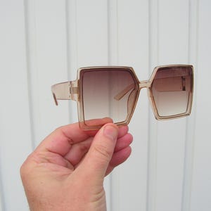 Puede incluir: Gafas de sol cuadradas de gran tamaño con un tinte marrón degradado. Las monturas son de color beige claro. Las gafas de sol están sujetas por una mano sobre un fondo blanco.