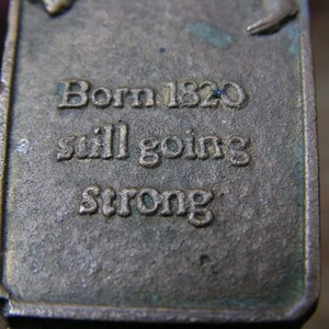May include: Close-up of a rectangular, aged metal object with the text "Born 1820 still going strong" embossed on its surface. The metal has a weathered appearance with hints of green oxidation.