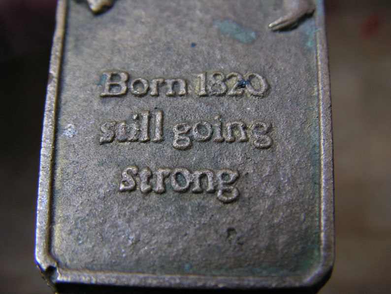 May include: Close-up of a rectangular, aged metal object with the text "Born 1820 still going strong" embossed on its surface. The metal has a weathered appearance with hints of green oxidation.