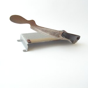 May include: A vintage metal paper cutter with a rusty blade and a black handle. The cutter has a white base with a beige cutting surface.