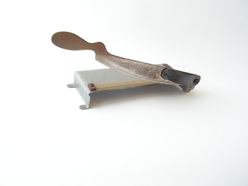 May include: A vintage metal paper cutter with a rusty blade and a black handle. The cutter has a white base with a beige cutting surface.