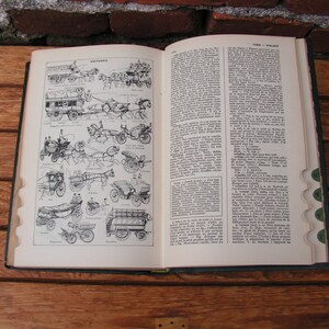 May include: An open vintage book with illustrations of horse-drawn carriages on the left page and text on the right. The book is on a wooden surface, with a brick wall in the background. The pages are aged, and the book has a dark green cover.