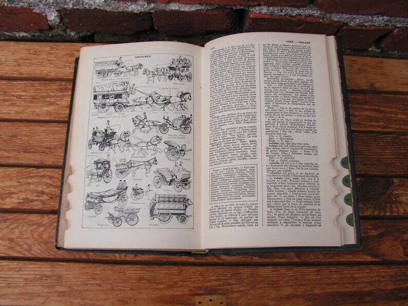 May include: An open vintage book with illustrations of horse-drawn carriages on the left page and text on the right. The book is on a wooden surface, with a brick wall in the background. The pages are aged, and the book has a dark green cover.