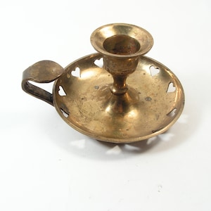May include: A brass candle holder with a heart-shaped design around the edge of the base. The holder has a handle and a single candle cup.