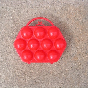 May include: Red plastic egg carrier with a handle. The carrier has 12 individual compartments for holding eggs.