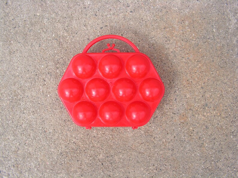 May include: Red plastic egg carrier with a handle. The carrier has 12 individual compartments for holding eggs.