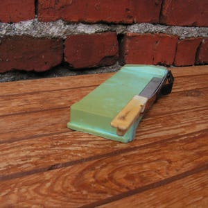 May include: A green and white plastic razor strop with a metal blade. The strop is sitting on a wooden surface.