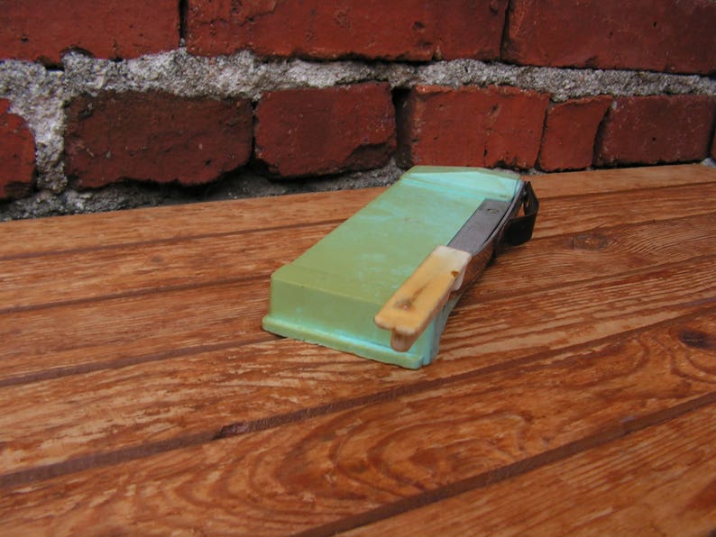 May include: A green and white plastic razor strop with a metal blade. The strop is sitting on a wooden surface.