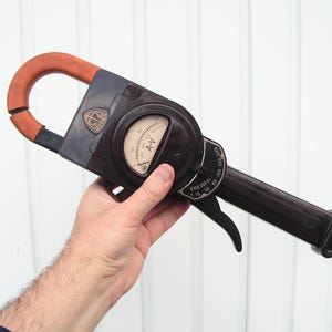 Puede incluir: Pinza amperimétrica eléctrica vintage negra y marrón con un mango naranja. El medidor tiene un dial circular con marcas A-V y un selector de rango. El dispositivo se sostiene en una mano.