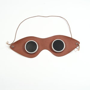 May include: A brown leather eye patch with two round black lenses. The eye patch has a strap that goes around the head.
