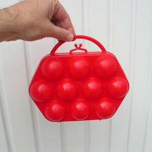 May include: A red plastic egg carrier with a handle. The carrier has 12 individual compartments for holding eggs.