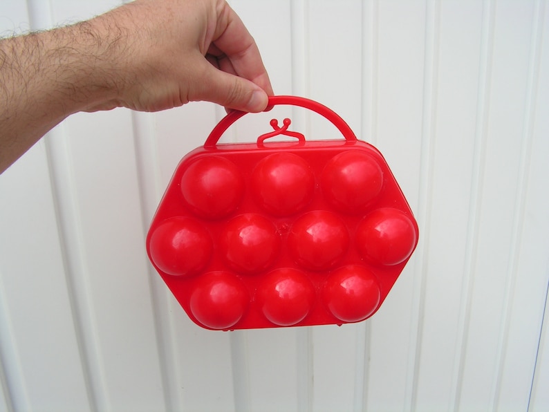 May include: A red plastic egg carrier with a handle. The carrier has 12 individual compartments for holding eggs.