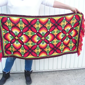 May include: A rectangular, hand-woven rug with a vibrant floral pattern. The design features red, yellow, and green geometric shapes. The rug has a red fringe along the edges.