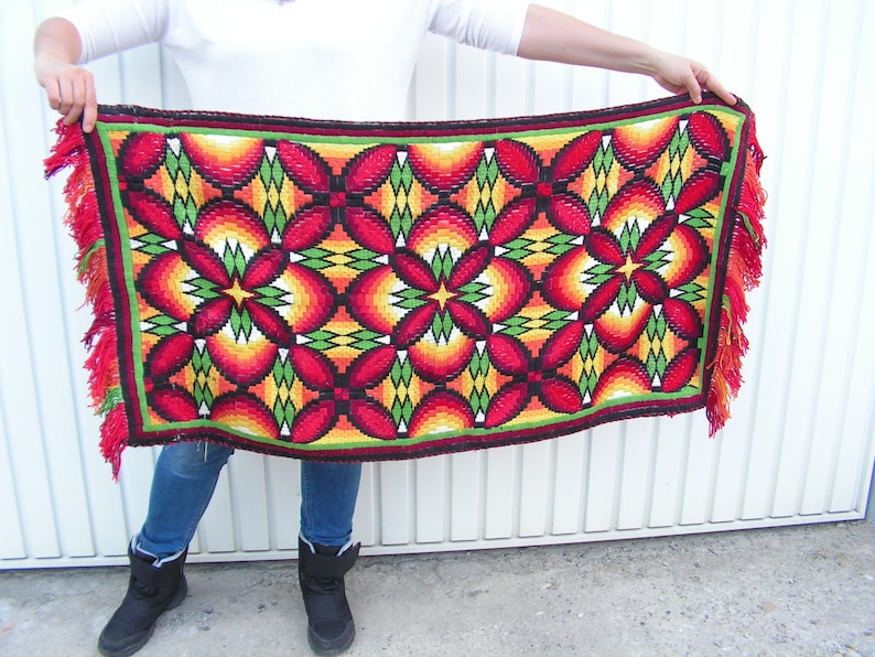 May include: A rectangular, hand-woven rug with a vibrant floral pattern. The design features red, yellow, and green geometric shapes. The rug has a red fringe along the edges.