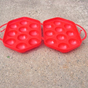 May include: Red plastic egg carton with a hinged lid and two handles. The carton has 12 individual compartments for holding eggs.