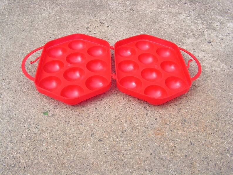 May include: Red plastic egg carton with a hinged lid and two handles. The carton has 12 individual compartments for holding eggs.